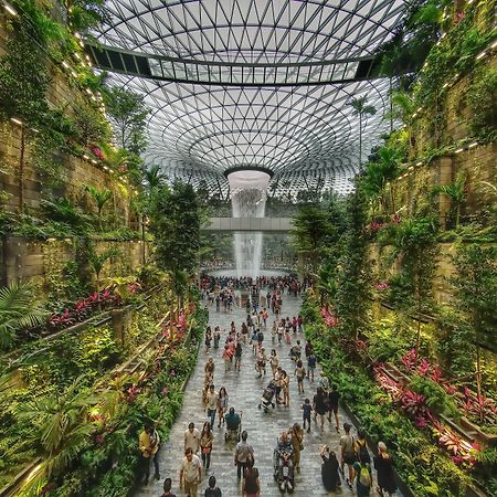 Crowne Plaza Changi Airport By Ihg Singapore Exterior photo The photo depicts a vibrant indoor garden space, characterized by lush greenery and a towering waterfall at its center. The setting features vertical gardens on stone walls, filled with various plants and flowers. People can be seen walking along the
