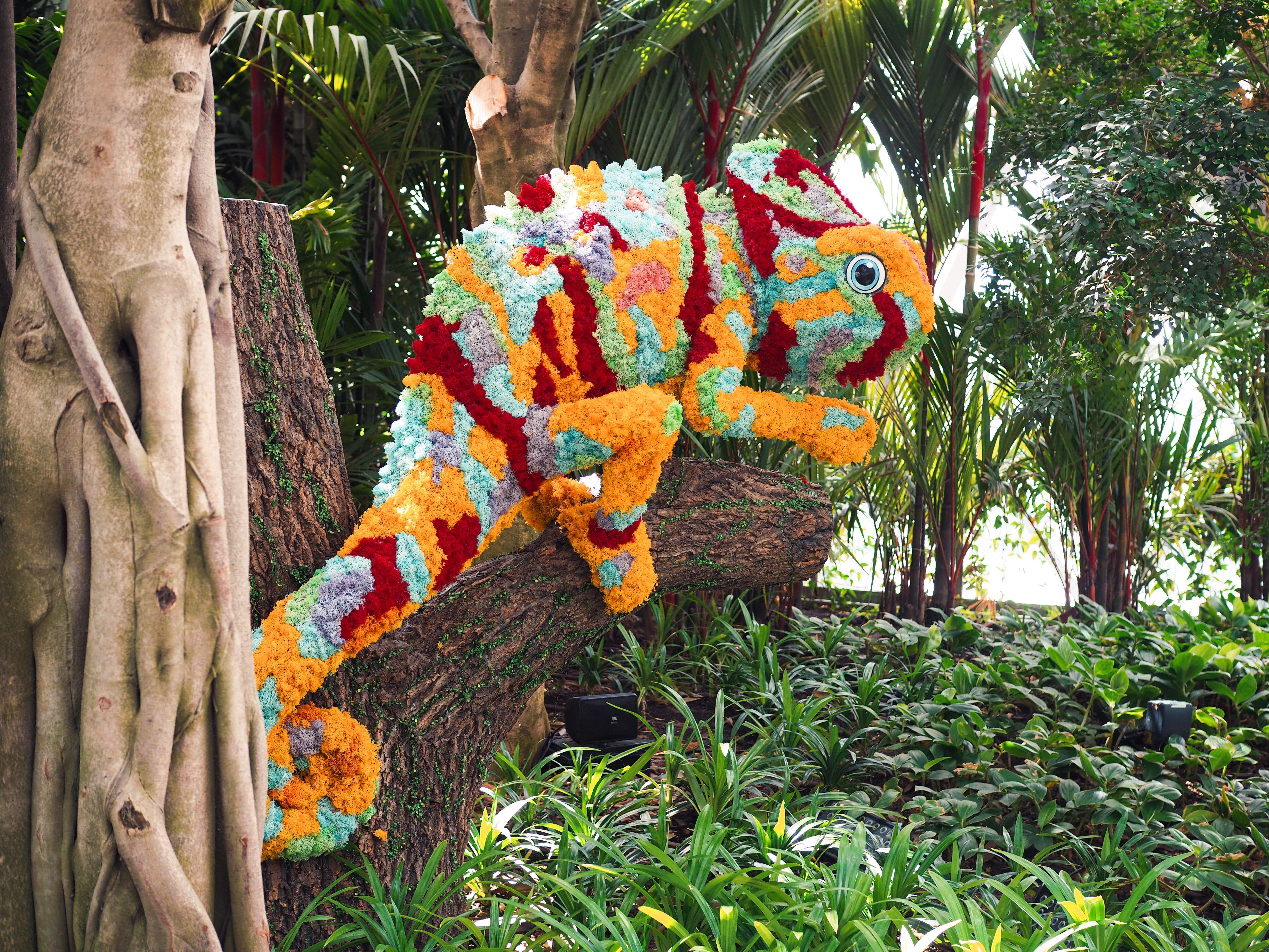 Crowne Plaza Changi Airport By Ihg Singapore Exterior photo The photo shows a vibrant and colorful chameleon sculpture made from flowers and plant materials. The chameleon is positioned on a tree trunk, blending in with the lush greenery of its surroundings. The colors of the sculpture include shades of orang