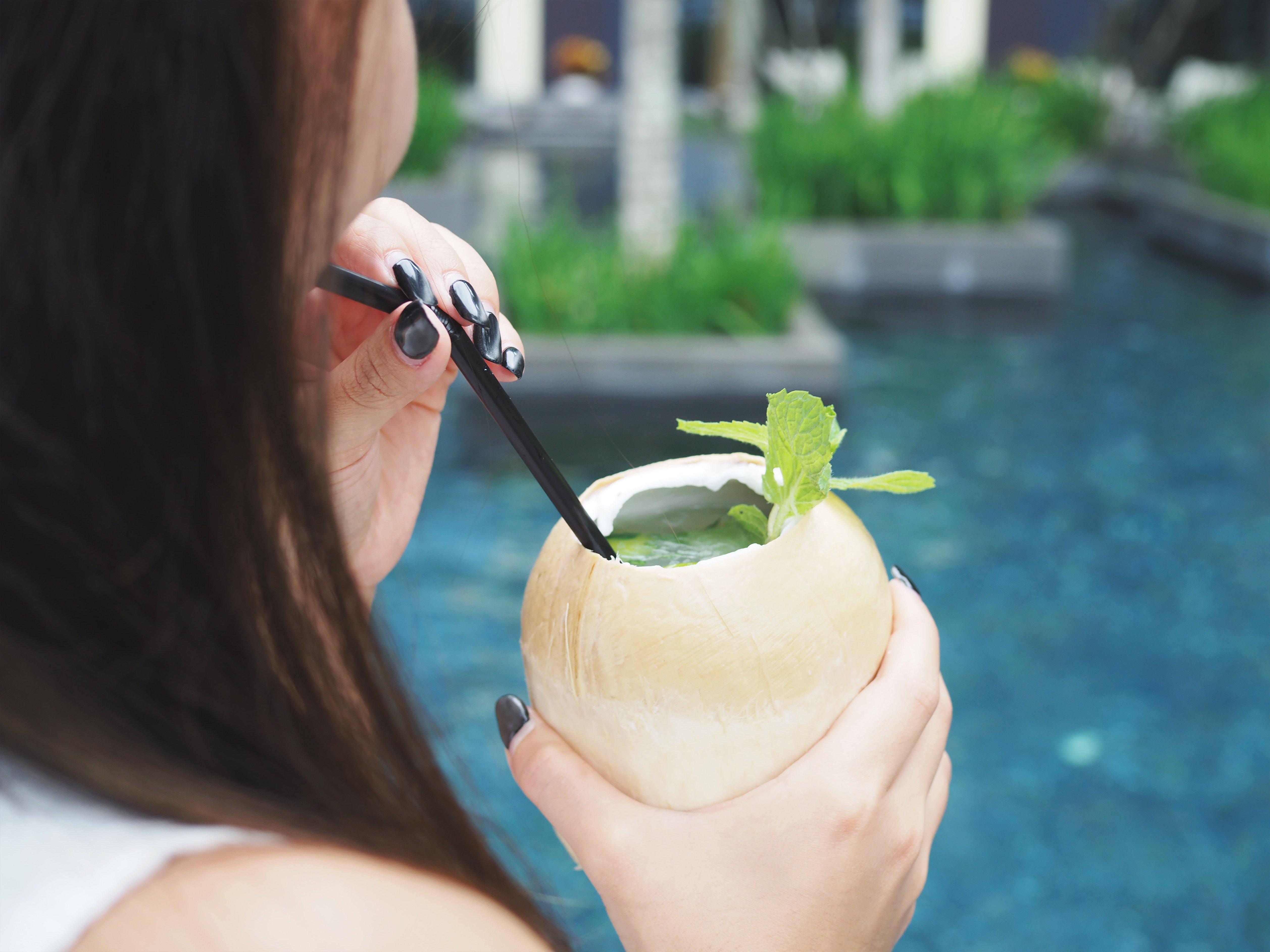 Crowne Plaza Changi Airport By Ihg Singapore Exterior photo The photo features a person holding a coconut drink with a straw. The drink is garnished with a sprig of mint. The background shows a blue pool, suggesting a tropical or relaxing setting. The focus is on the drink and the person's hand.