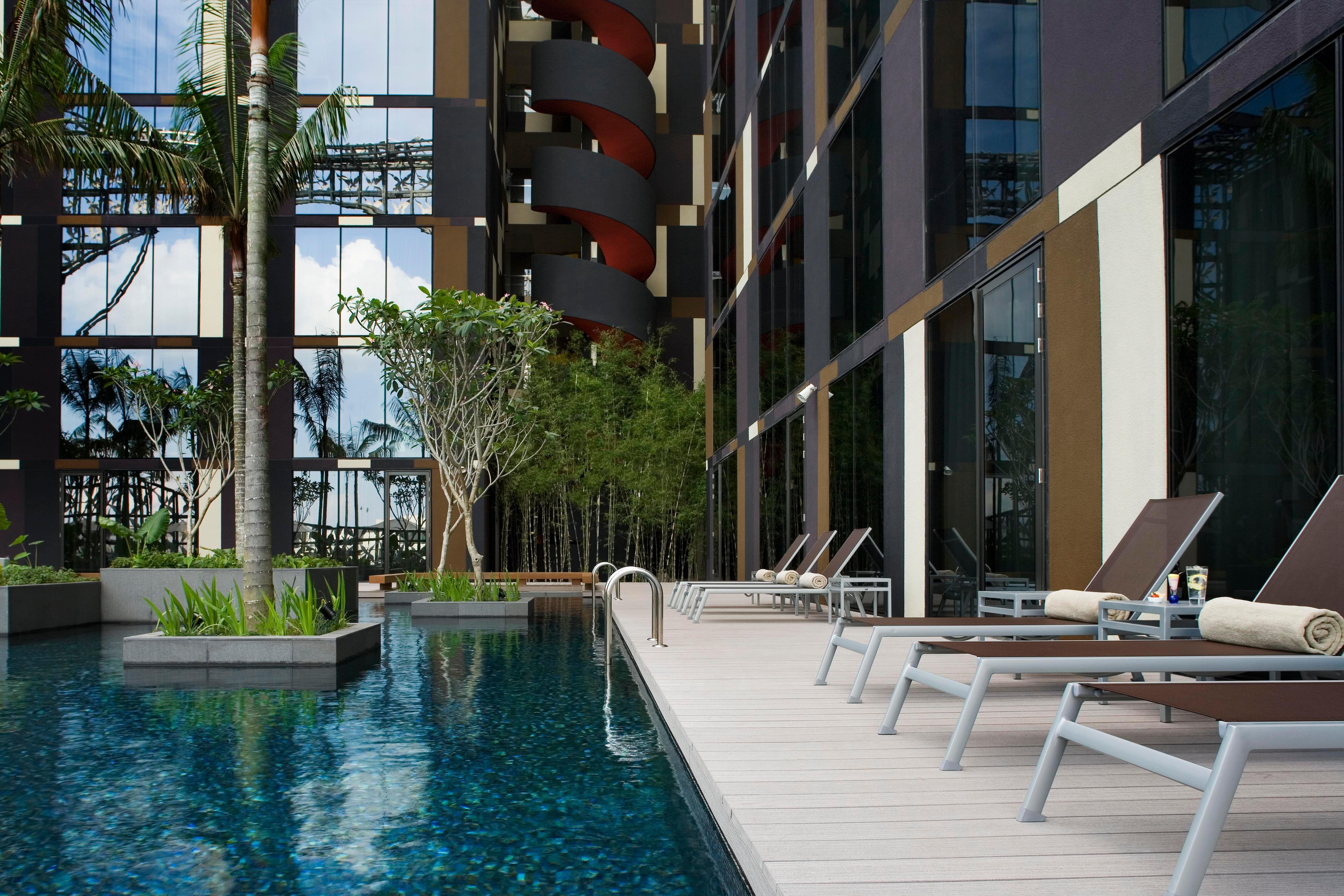 Crowne Plaza Changi Airport By Ihg Singapore Facilities photo The photo shows a modern swimming pool area adjacent to a building. The pool is surrounded by sleek wooden decking and features a clear blue water surface. There are several lounge chairs positioned around the pool for relaxation. Lush greenery, incl
