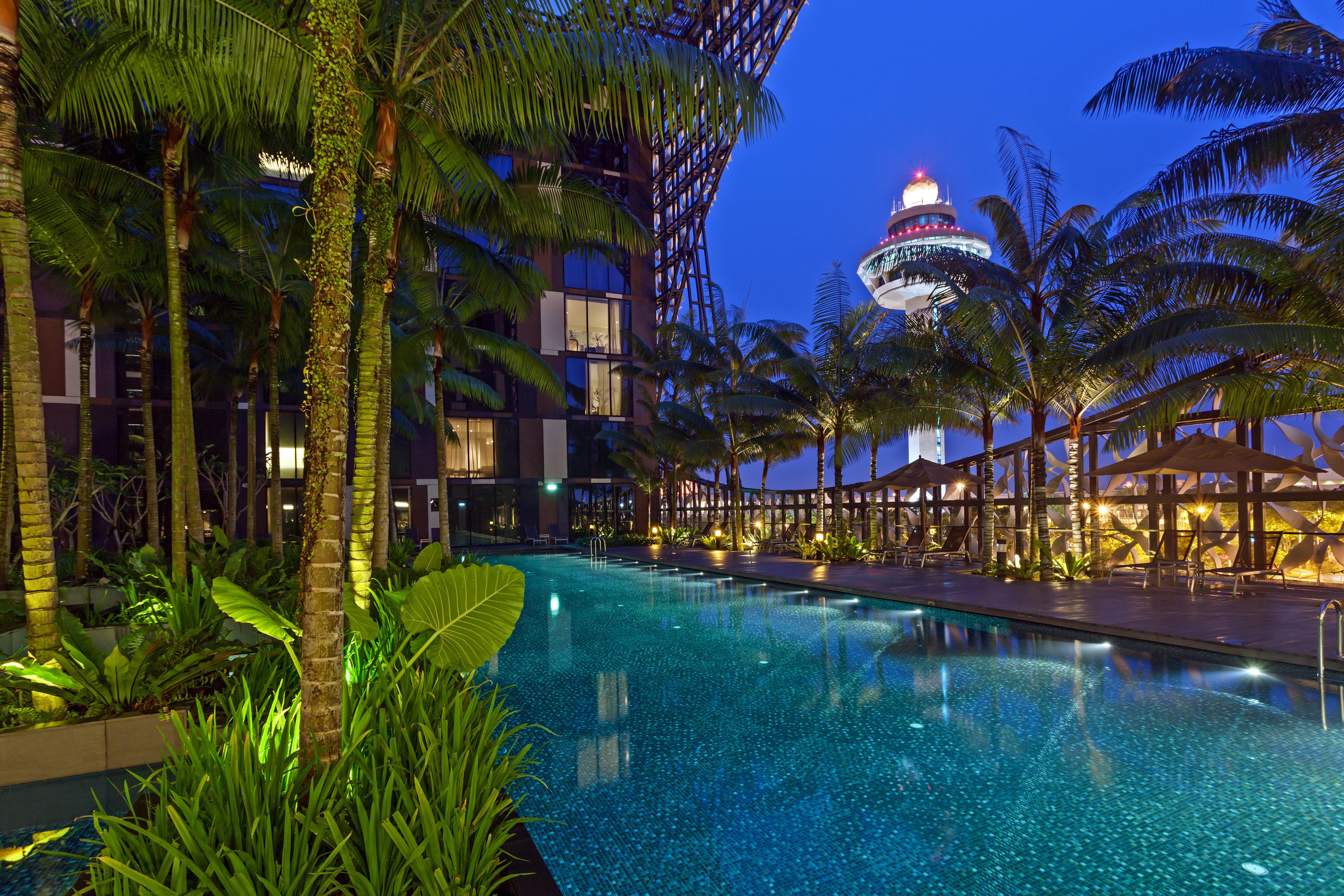 Crowne Plaza Changi Airport By Ihg Singapore Exterior photo The photo depicts a luxurious outdoor swimming pool surrounded by lush palm trees and vibrant greenery. The scene is set during the evening, with soft lighting illuminating the water and plants. In the background, you can see a modern building struct