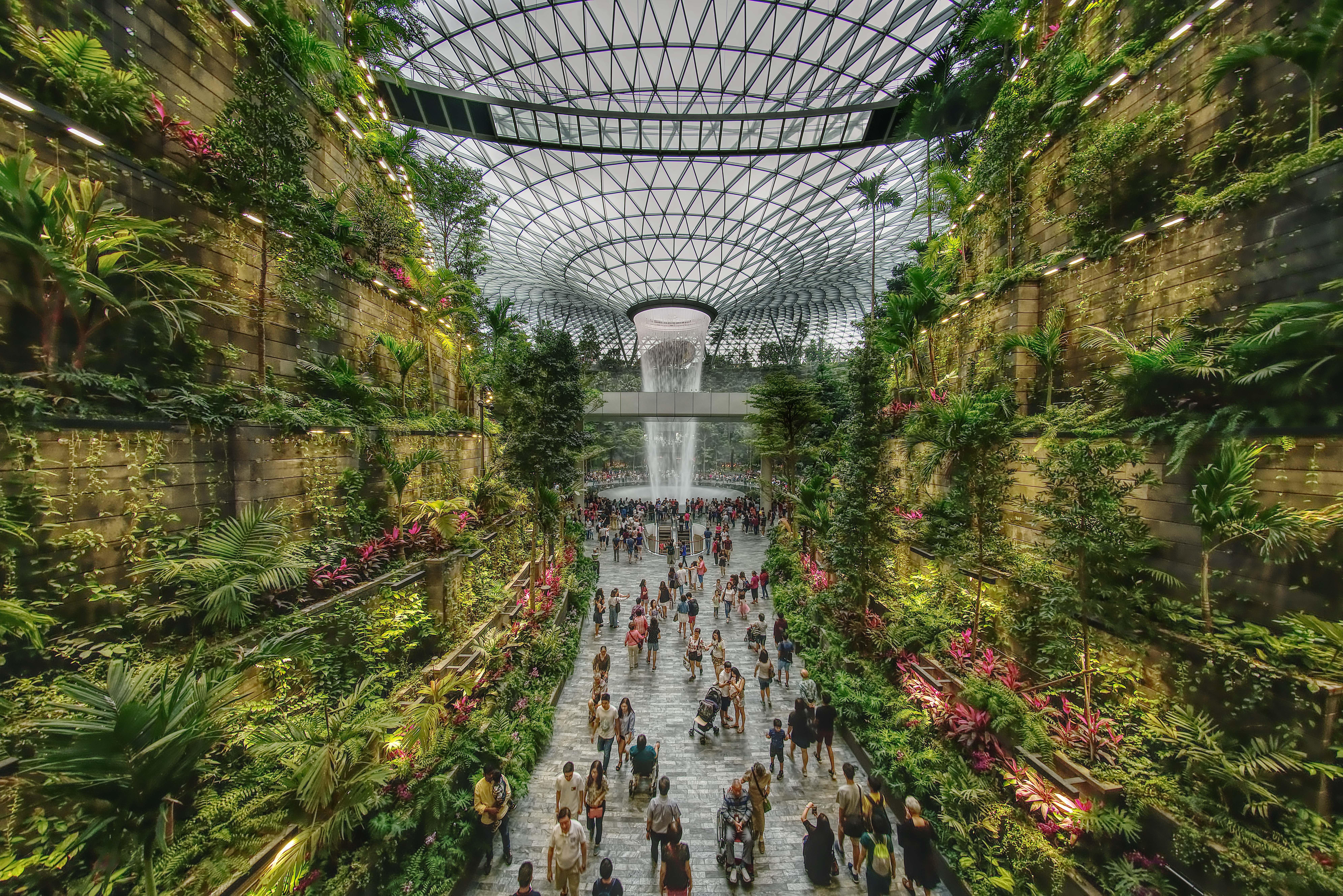 Crowne Plaza Changi Airport By Ihg Singapore Exterior photo The photo depicts a vibrant indoor garden space, characterized by lush greenery and a towering waterfall at its center. The setting features vertical gardens on stone walls, filled with various plants and flowers. People can be seen walking along the