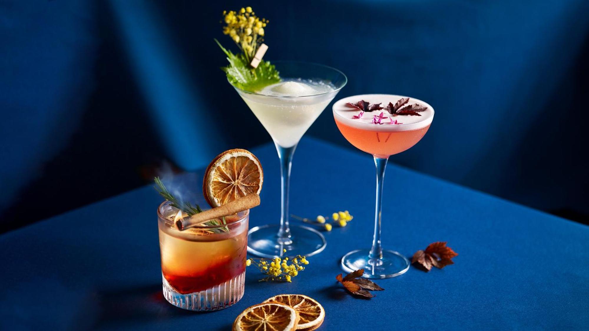 Crowne Plaza Changi Airport By Ihg Singapore Exterior photo The photo shows a stylish arrangement of three cocktails on a blue surface. 

1. **Left Cocktail**: A lowball glass containing a drink with a reddish hue, garnished with a dried orange slice and a cinnamon stick. 

2. **Middle Cocktail**: A classic m