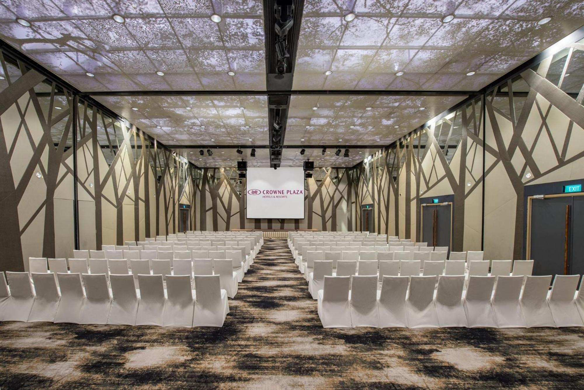 Crowne Plaza Changi Airport By Ihg Singapore Exterior photo The photo shows a modern conference or event space. The room features rows of white chairs arranged in a theater-style layout, facing a large screen with a simple logo. The ceiling is designed to resemble a natural setting, with images that suggest t