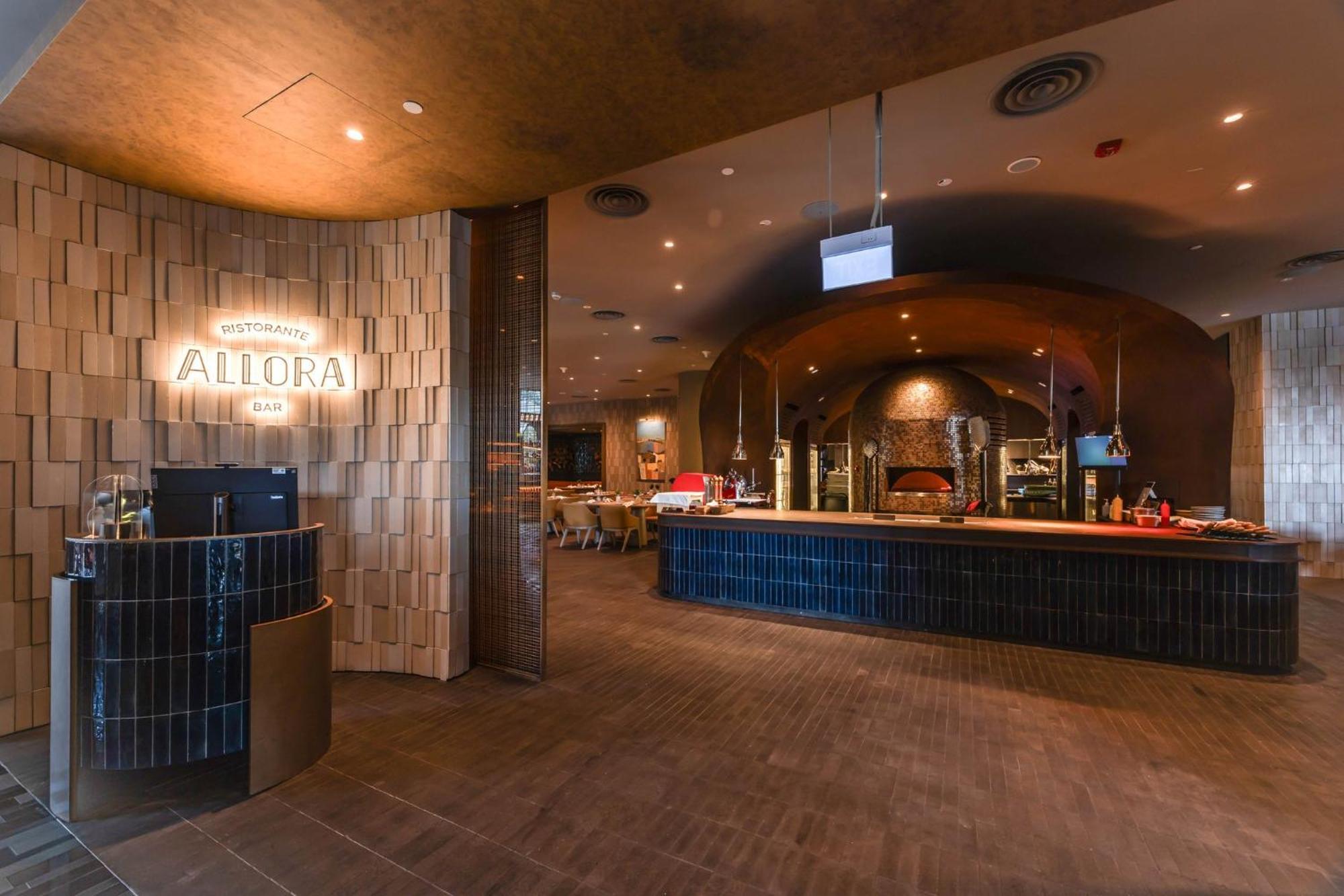 Crowne Plaza Changi Airport By Ihg Singapore Exterior photo The photo shows the interior of a restaurant named "Allora." The design features a contemporary style with a warm color palette. In the foreground, there is a curved bar area with a sign indicating the restaurant's name. The walls are adorned with te