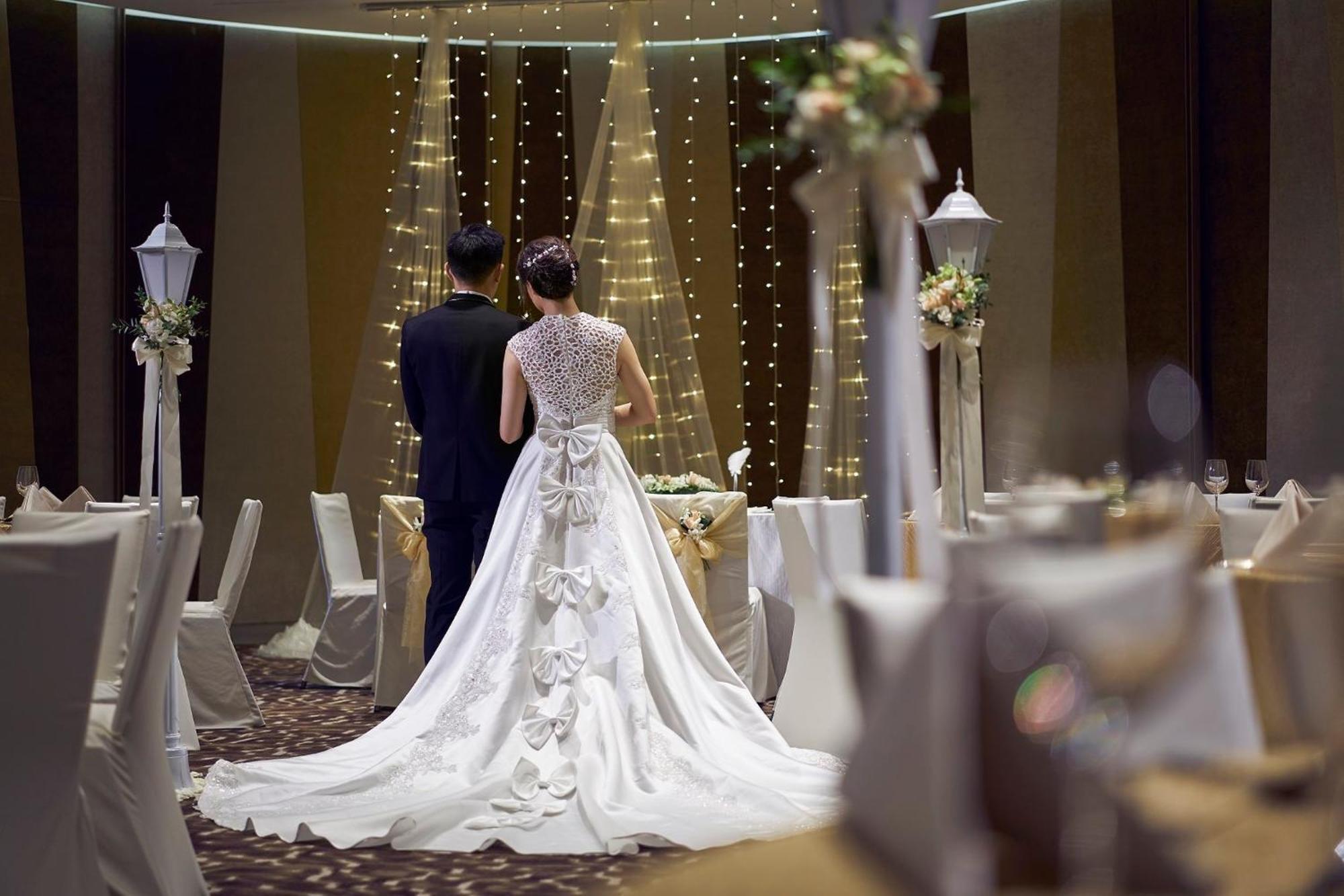 Crowne Plaza Changi Airport By Ihg Singapore Exterior photo The photo features a couple at a wedding venue. The bride, wearing an elegant white gown with intricate details and a long train, is seen from behind. The groom stands beside her, dressed in a dark suit. The background showcases a beautifully decorat