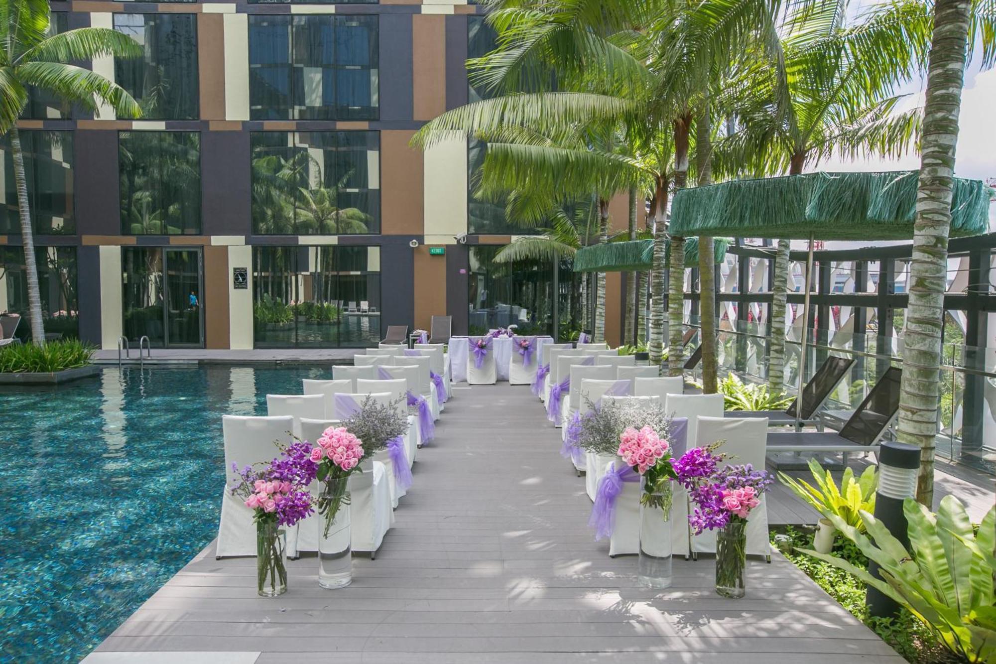 Crowne Plaza Changi Airport By Ihg Singapore Exterior photo The photo shows a beautifully arranged outdoor setting for a wedding or a similar event. There is a long aisle made of wooden planks leading to a pool area, flanked by neatly arranged chairs. Each chair is adorned with purple ribbons and decorated wi