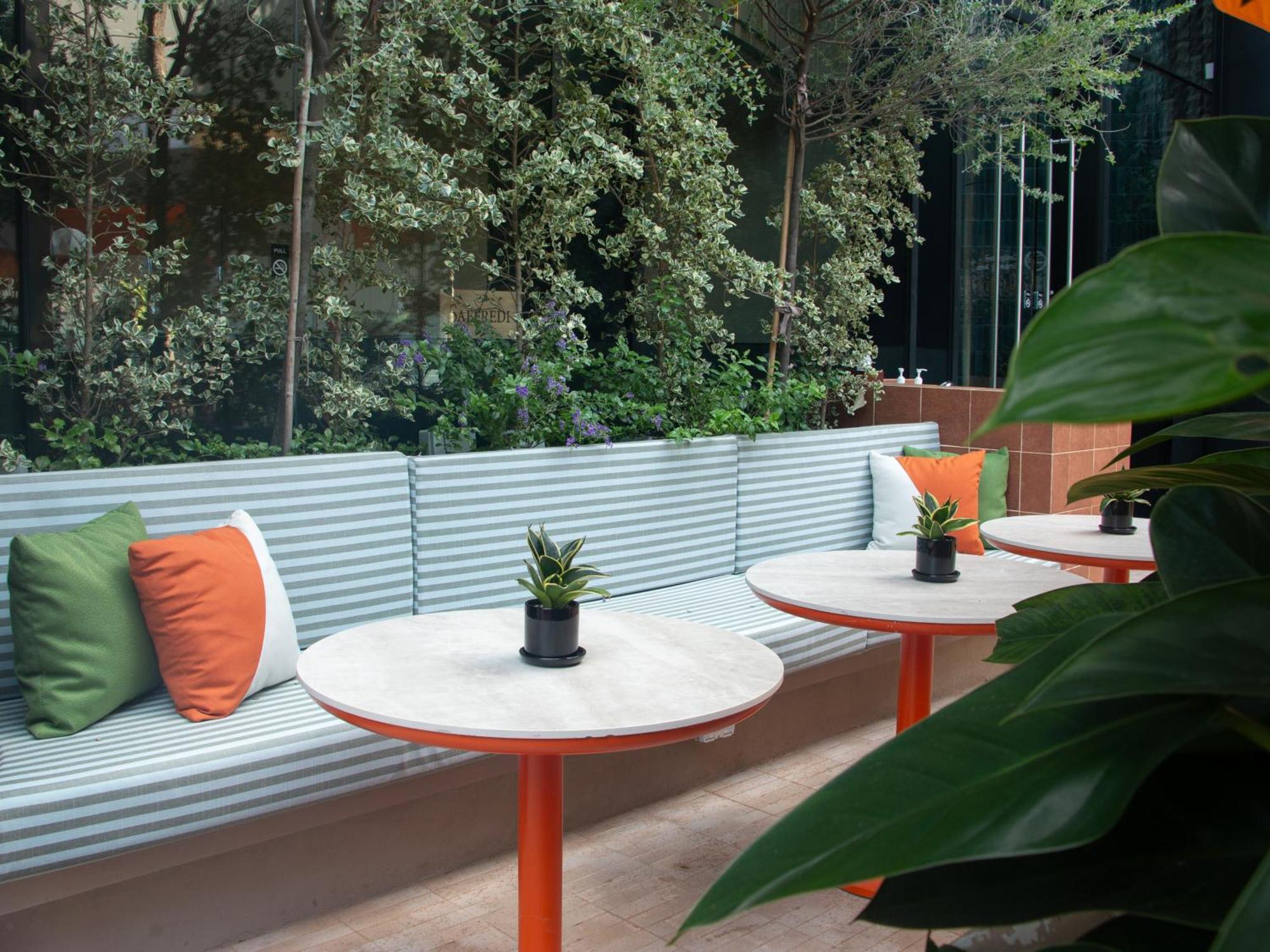 Crowne Plaza Changi Airport By Ihg Singapore Exterior photo The photo shows a modern outdoor seating area. There is a long bench with a corrugated metal backrest, decorated with colorful cushions in green and orange. In front of the bench, there are circular tables with white marble tops and orange legs. Each