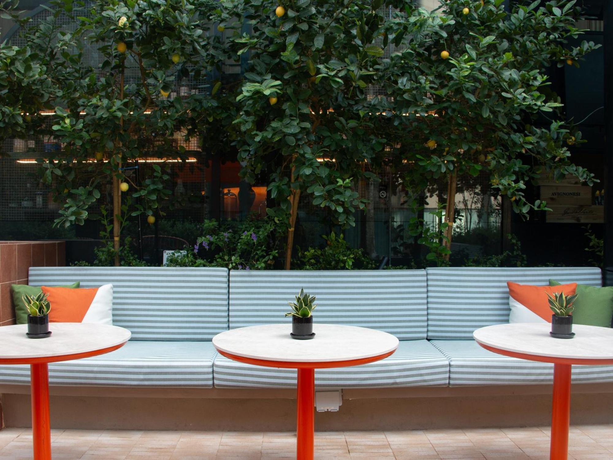 Crowne Plaza Changi Airport By Ihg Singapore Exterior photo The photo shows a cozy seating area featuring a long, light blue bench with colorful cushions in orange and green. In front of the bench, there are three round tables with bright red bases and small plants in black pots on each table. The background 