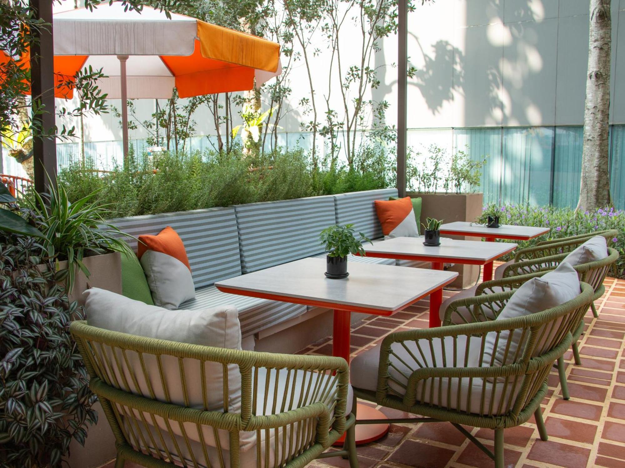 Crowne Plaza Changi Airport By Ihg Singapore Exterior photo The photo shows a cozy outdoor seating area. There are several tables with red metal legs, each accompanied by comfortable chairs. The chairs have a woven design and are in shades of green. There are cushions in various colors on the chairs for added
