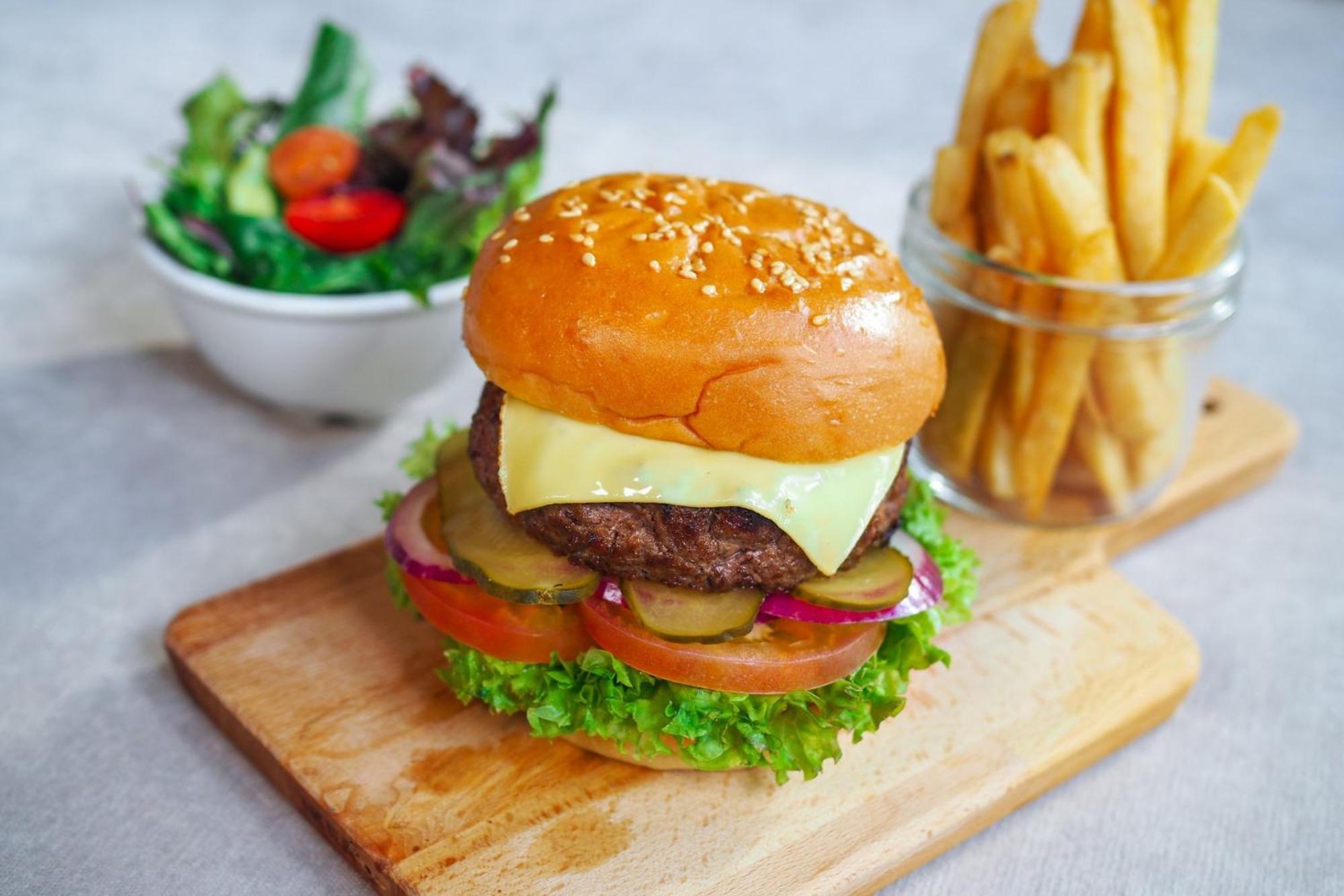 Crowne Plaza Changi Airport By Ihg Singapore Exterior photo The photo shows a delicious looking hamburger with a sesame seed bun. The burger is layered with a beef patty, melted cheese, lettuce, tomato slices, pickles, and red onion. It is presented on a wooden cutting board. Next to the burger, there is a sm