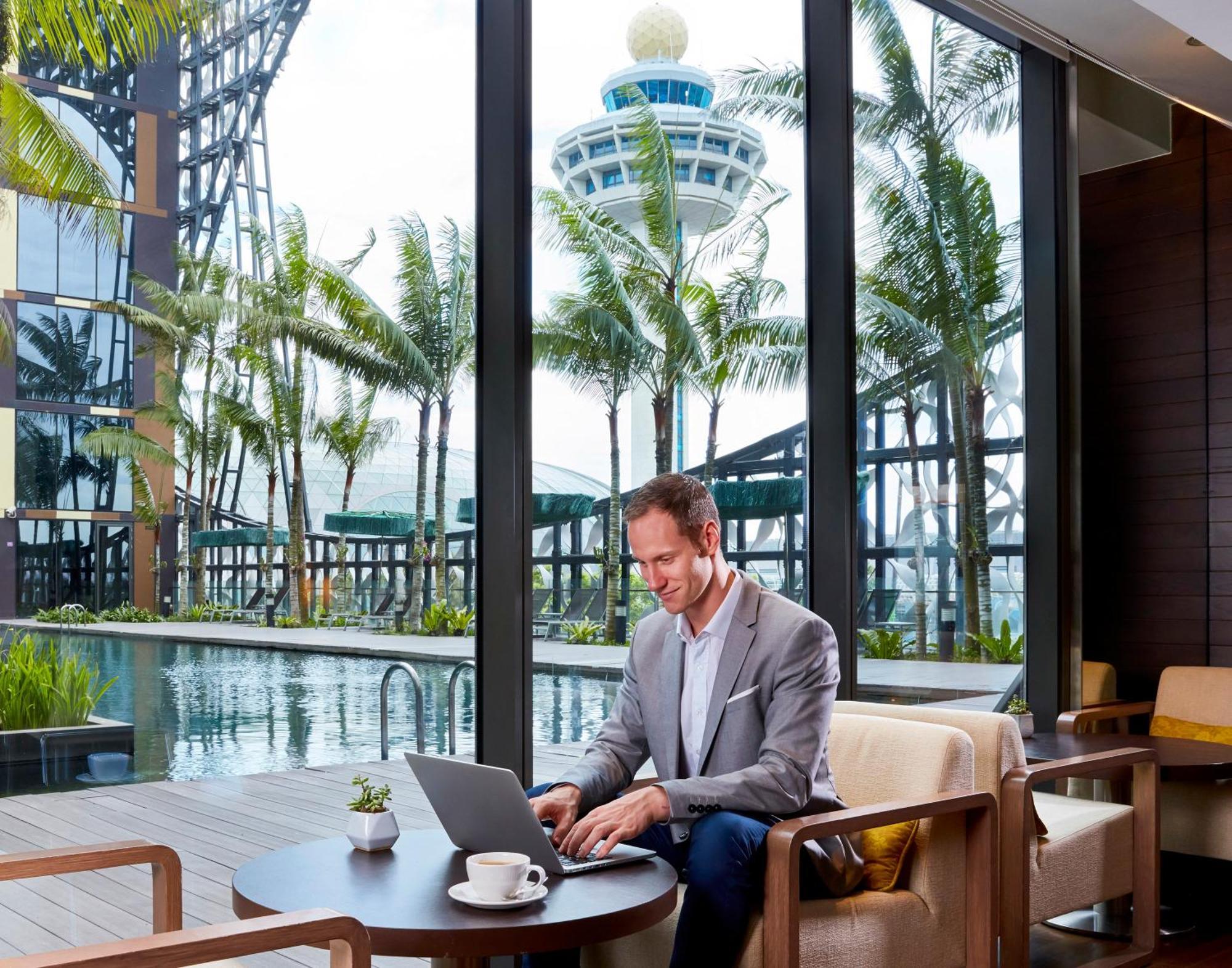 Crowne Plaza Changi Airport By Ihg Singapore Exterior photo The photo depicts a man sitting at a modern table working on a laptop. He is dressed in a smart, light gray blazer and appears focused on his task. The setting has large windows that offer a view of palm trees and a pool, suggesting a luxurious and t