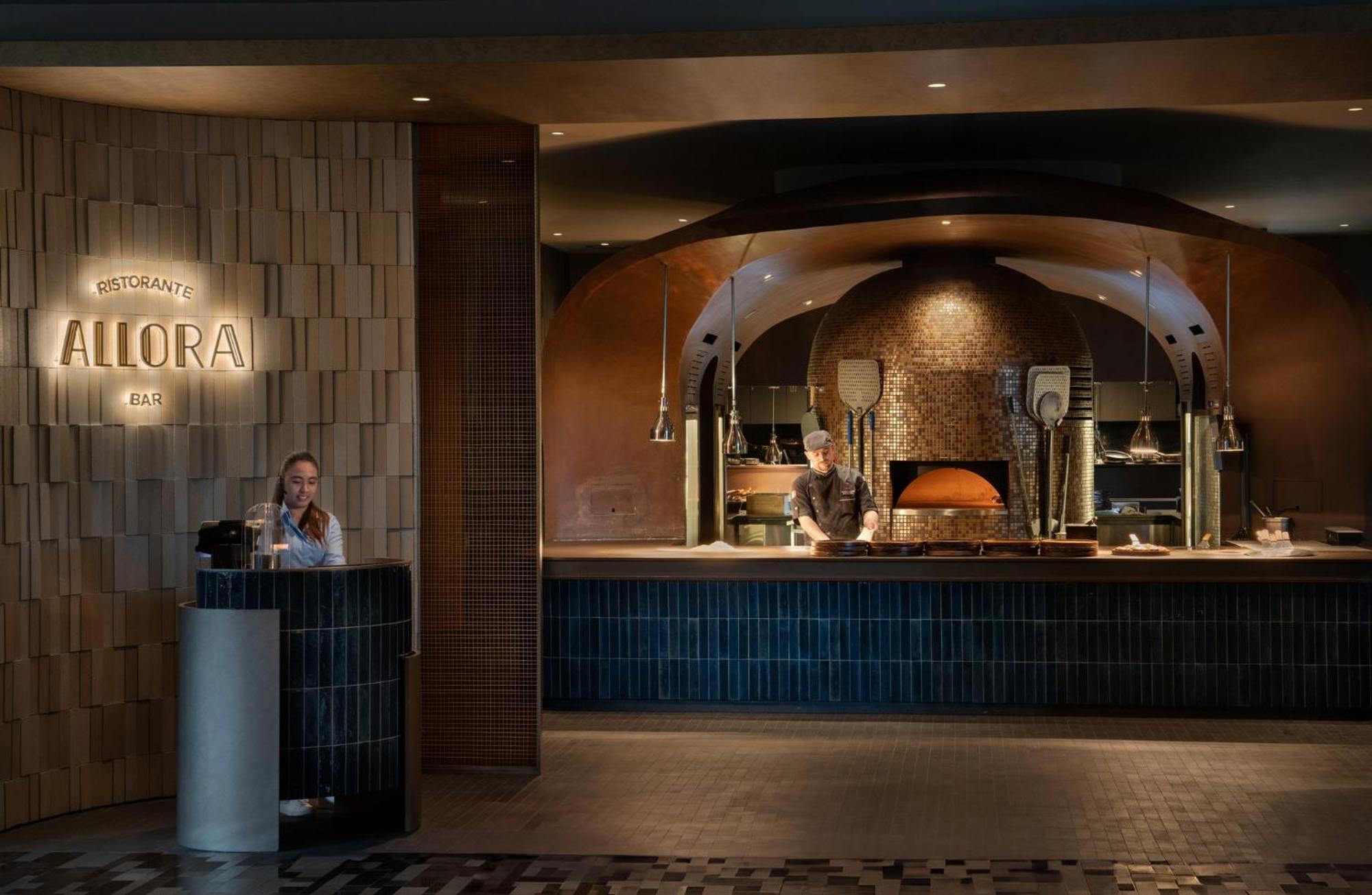 Crowne Plaza Changi Airport By Ihg Singapore Exterior photo The photo showcases a restaurant interior, featuring a modern and stylish design. On the left side, there’s a reception area with a hostess. The central part of the image displays an open kitchen with two cooks; one is visible behind a counter, and t