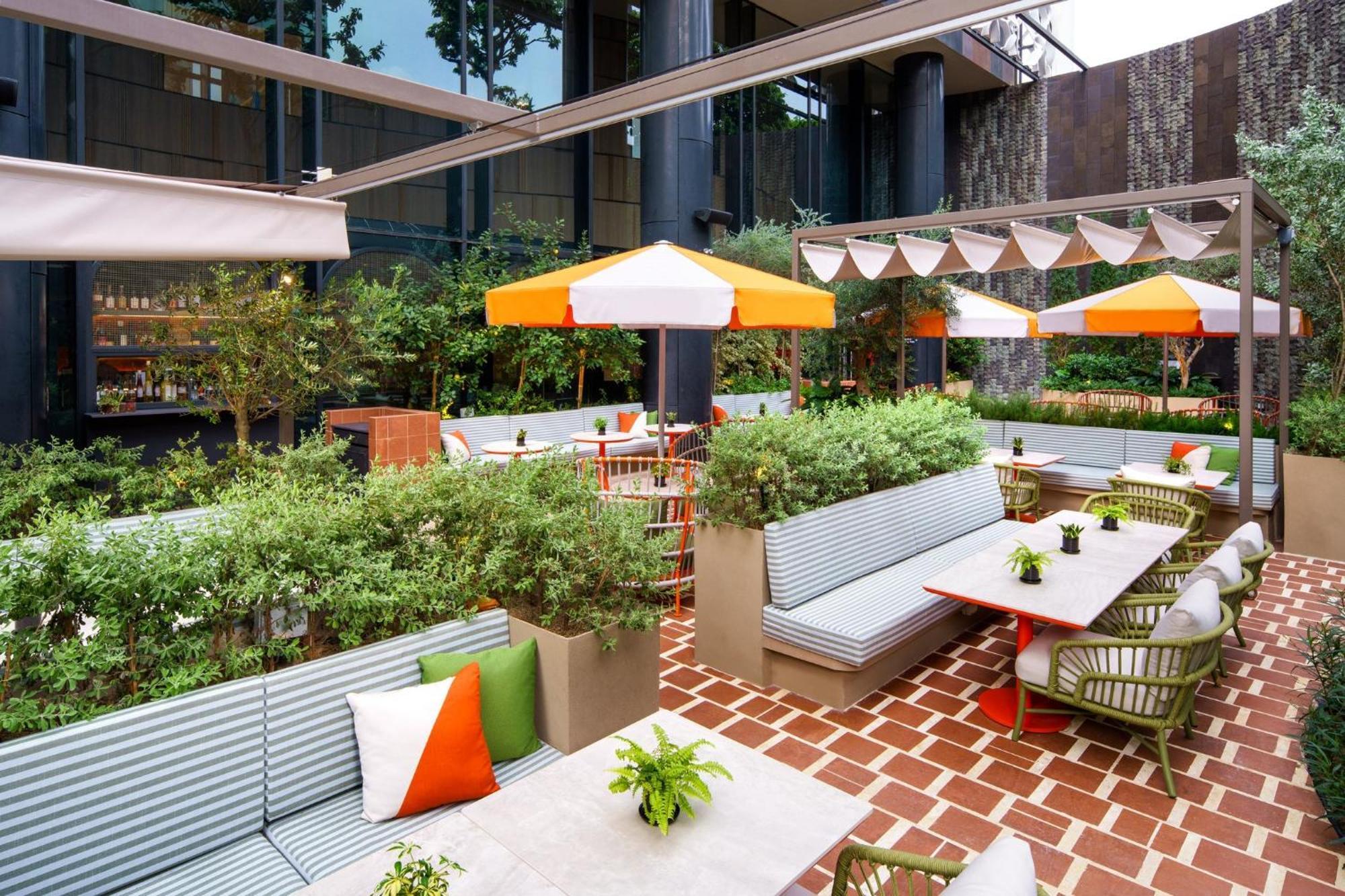 Crowne Plaza Changi Airport By Ihg Singapore Exterior photo The photo depicts a stylish outdoor seating area in a modern setting. It features several light gray benches with colorful cushions, arranged around tables. The space is adorned with green plants and small potted flowers, adding a touch of nature. Th