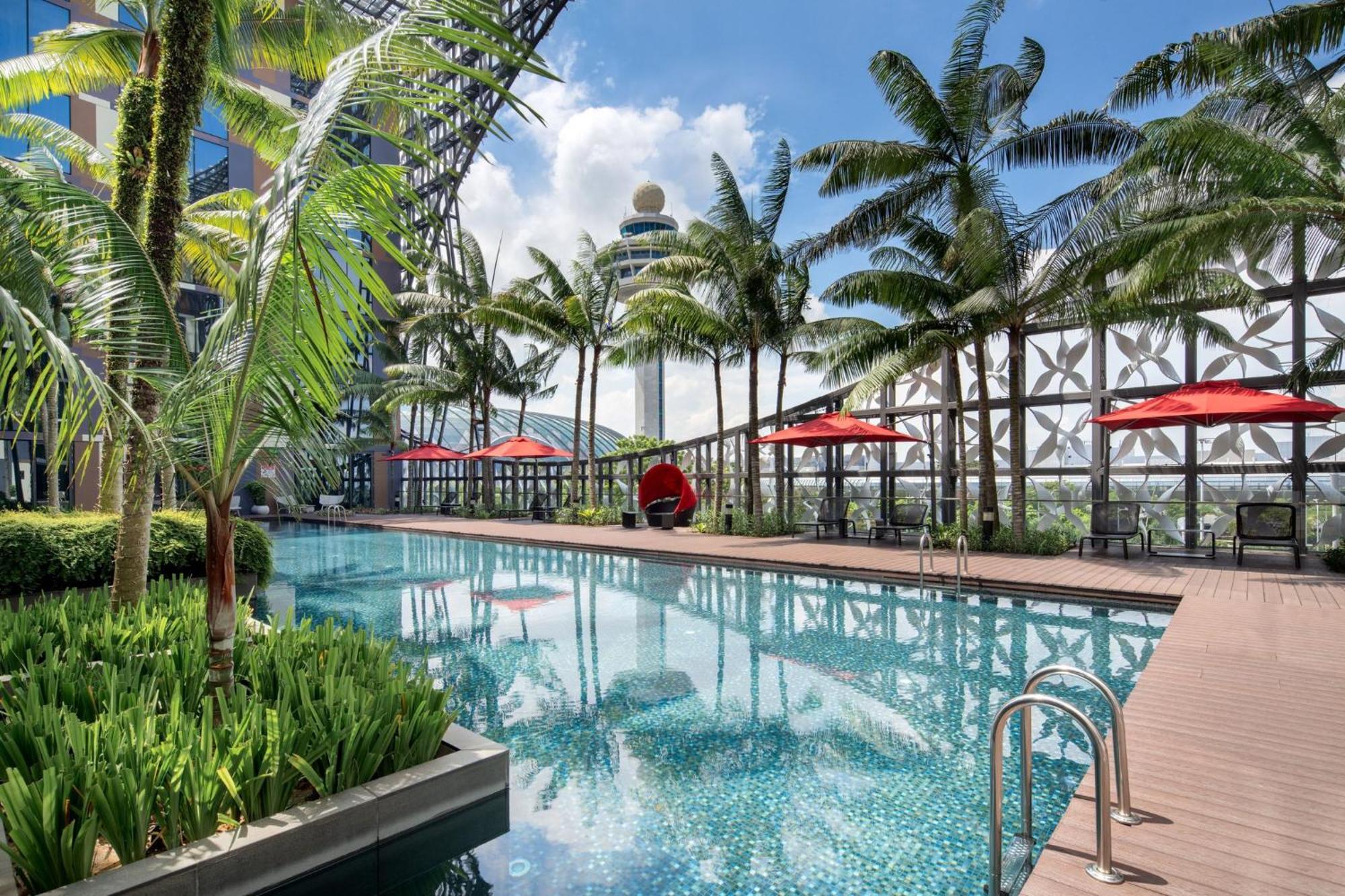 Crowne Plaza Changi Airport By Ihg Singapore Exterior photo The image shows a luxurious pool area surrounded by palm trees. The pool has a clear blue water surface, with small tiles reflecting light. On one side, there are lounge chairs with red umbrellas providing shade. There are also some decorative plants